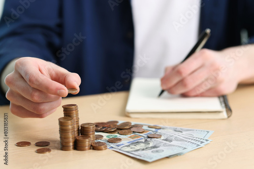 Financial savings. Man stacking coins while writing down notes at wooden table, closeup