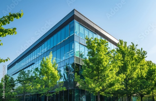 A sleek, modern office building with large glass windows and green trees outside, symbolizing eco-friendly business practices. The sky is a clear blue in the background