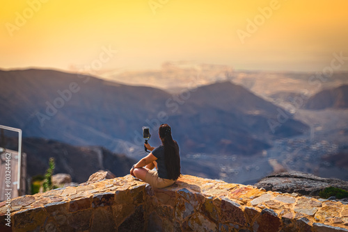 Young woman with long hair and sporty outfit sitting on the ground with arms wide open, looking at the mountainous scenic view ahead. Roads can be seen on the background.          photo
