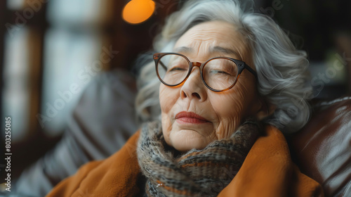 An elderly woman is sitting in a chair with her eyes closed. She is wearing a brown coat and a scarf. Her hair is gray and she is wearing glasses.