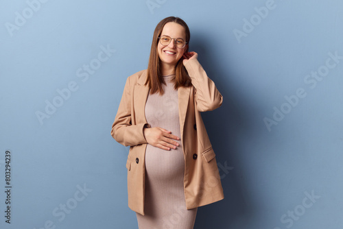 Attractive optimistic positive pregnant woman in beautiful dress and jacket posing isolated over blue background looking at camera expressing love and tenderness