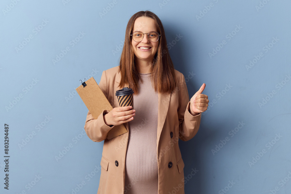 Cheerful pregnant woman in beautiful dress and jacket posing isolated over blue background showing like gesture thumb up enjoying her work holding coffee and clipboard Cheerful pregnant woman in beautiful dress and jacket posing isolated over blue background showing like gesture thumb up enjoying her work holding coffee and clipboard