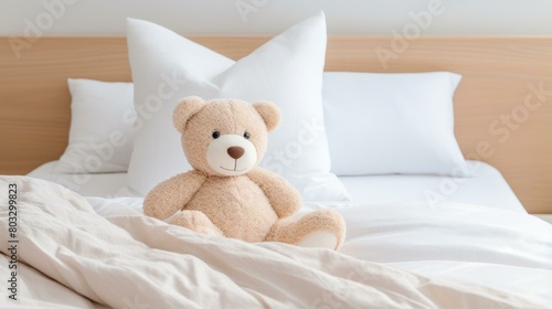 A cute teddy bear sitting on a bed with white pillows and a white blanket