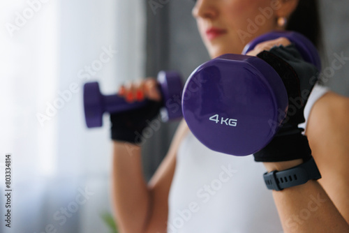 Watch as the woman holds two purple dumbbells in her hands, wearing sportswear and personal protective equipment. Her arm muscles tense with each gesture, engaging her chest and thigh muscles