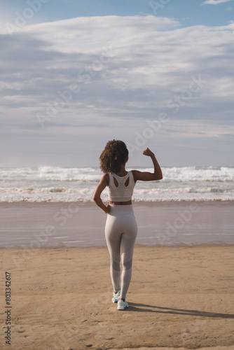 Back view of fit black woman flexing arm towards the sea at the beach. Fitness motivation and success concept. Successful sporty female exercising outdoor.