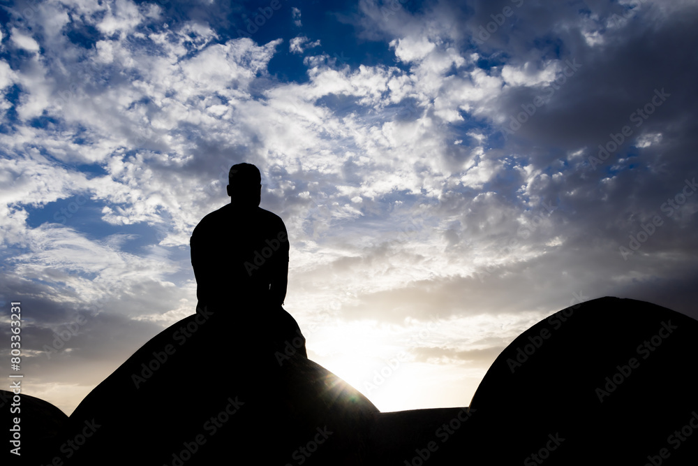 backlit shot of isolated man sitting at wall watching sunset with dramatic sky at dusk backlit shot of isolated man sitting at wall watching sunset with dramatic sky at dusk