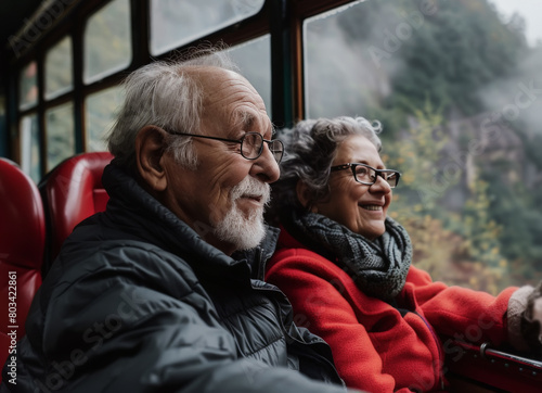 Hombre y mujer senior de más de 70 años con gafas graduadas, viajando sentados juntos y sonrientes en un tren antiguo de madera , sobre fondo desenfocado de paisaje verde montañoso photo