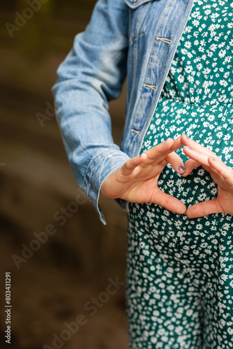 woman in green floral dress holding her hands in a heart shape over her pregnant belly