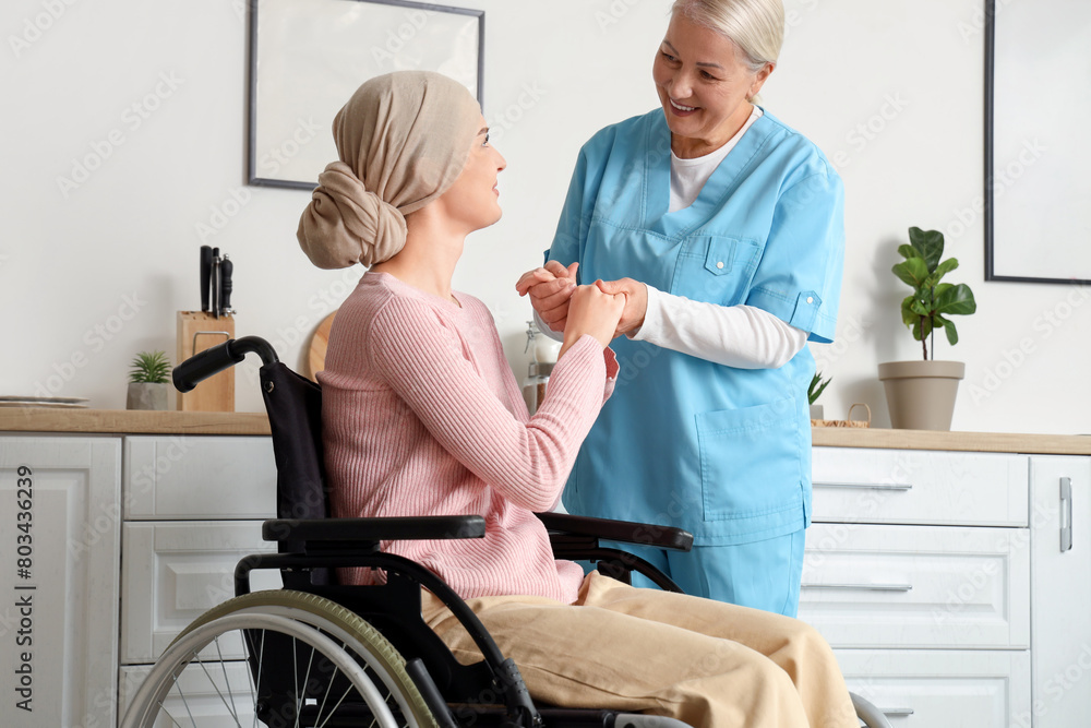 Fototapeta premium Young woman after chemotherapy in wheelchair with nurse holding hands at home. Stomach cancer concept