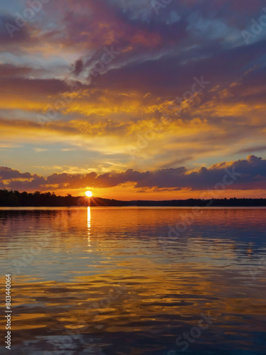 Illustrate the mesmerizing sight of a stunning sunset painting the horizon above a calm lake, its vibrant colors mirrored in the shimmering water below.