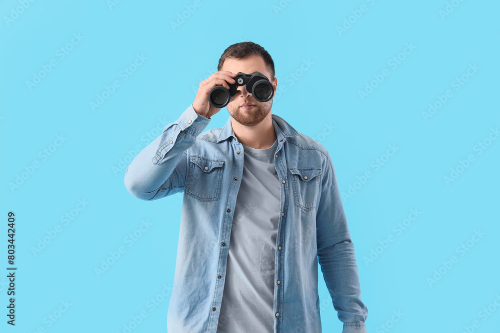 Young man looking through binoculars on blue background Young man looking through binoculars on blue background
