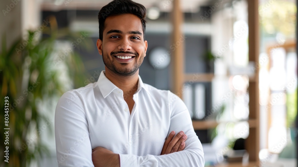 Obraz premium Smiling confident young Indian businessman in white shirt standing with crossed arms the background of blurred office