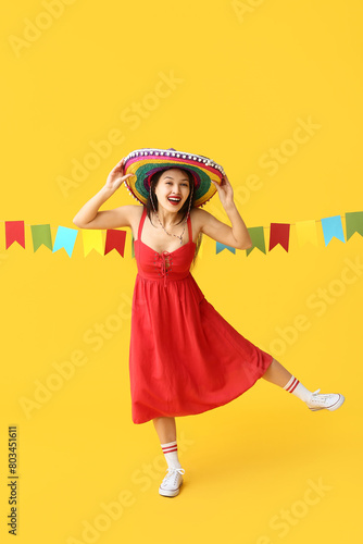 Happy young woman in Mexican sombrero hat and with garland on yellow background