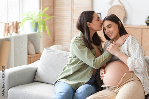 Young lesbian woman kissing her pregnant wife on sofa at home