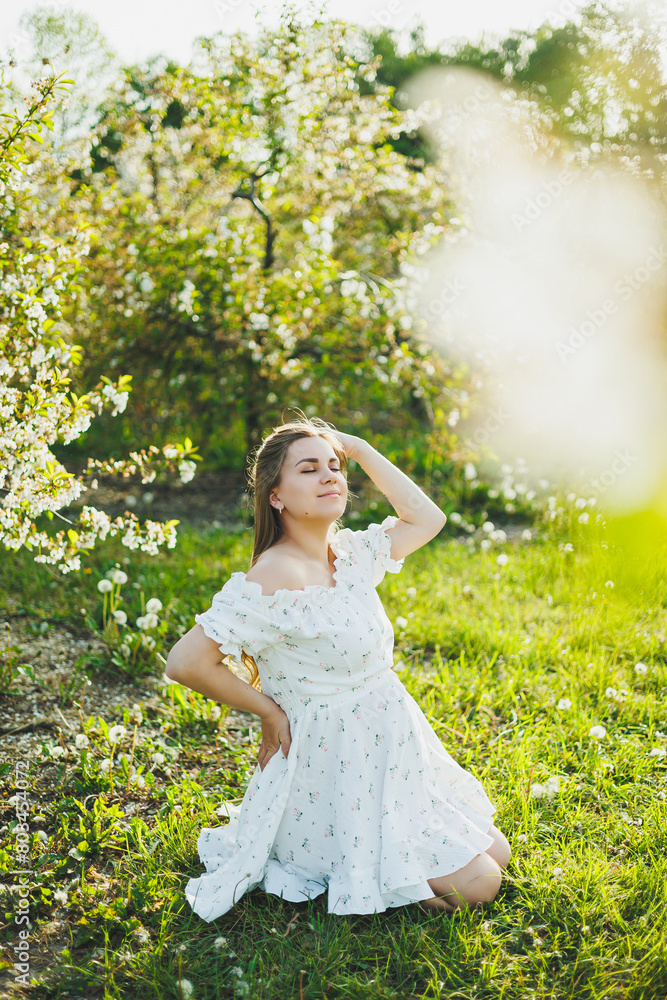 A happy pregnant woman in a summer white muslin dress is sitting on the grass in a blooming garden. Future mother, a woman expecting a baby. Pregnancy period A happy pregnant woman in a summer white muslin dress is sitting on the grass in a blooming garden. Future mother, a woman expecting a baby. Pregnancy period