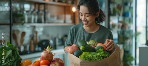 In her thirties, an Asian woman joyfully unpacks a paper bag filled with groceries onto a white table, showcasing a variety of fruits and vegetables. photo