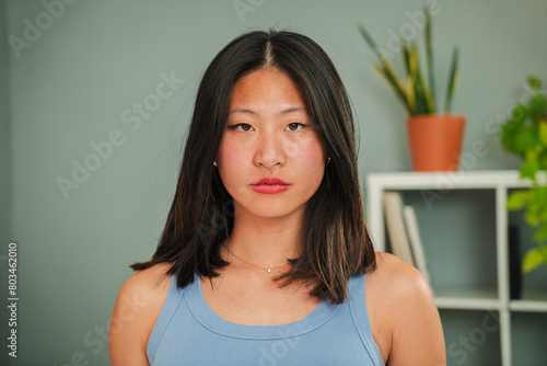 Close up individual portrait of serious asian young woman looking at camera standing at home linving room. One thoughtufl chinese female with confident, pensive and moody expression Real girl frowning photo