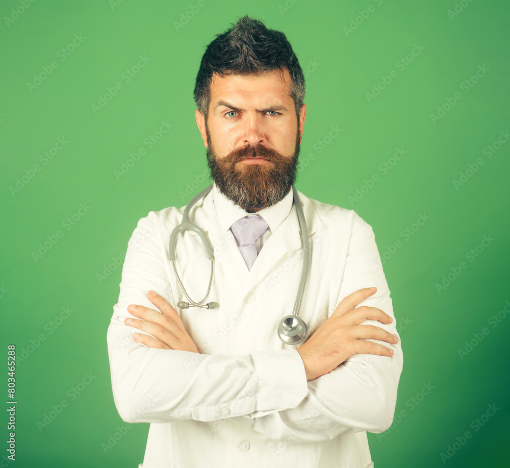 Profession, medicine, healthcare and medical staff concept. Handsome bearded intern, physician in white uniform with crossed arms in clinic. Serious male doctor in workwear with stethoscope on neck. Profession, medicine, healthcare and medical staff concept. Handsome bearded intern, physician in white uniform with crossed arms in clinic. Serious male doctor in workwear with stethoscope on neck.