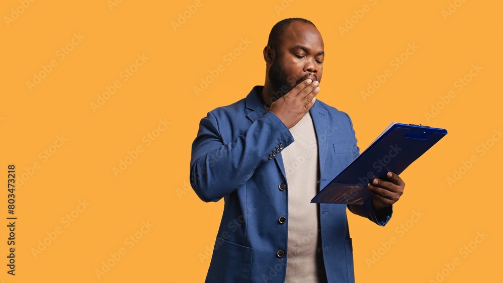 Man browsing though financial audit report on clipboard, yawning, feeling tired, studio background. Bored african american worker exhausted while reading paperwork on notepad, camera B Man browsing though financial audit report on clipboard, yawning, feeling tired, studio background. Bored african american worker exhausted while reading paperwork on notepad, camera B