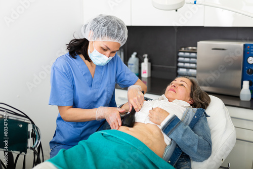 Confident female doctor performing radiofrequency lifting procedure on abdomen of mature woman