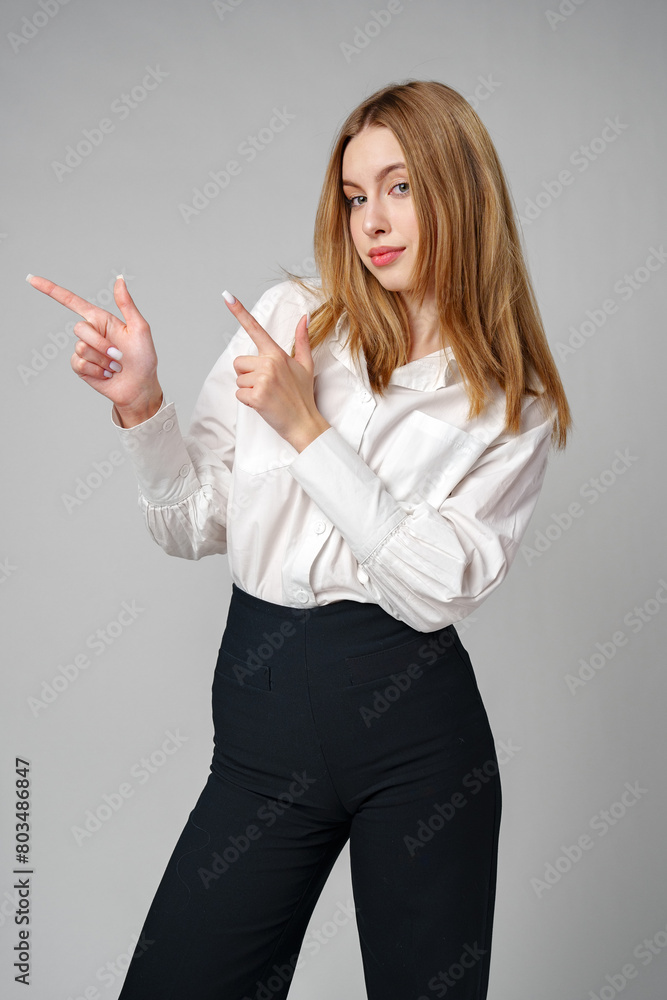 Young blonde woman in formal outfit pointing to the side against gray background Young blonde woman in formal outfit pointing to the side against gray background