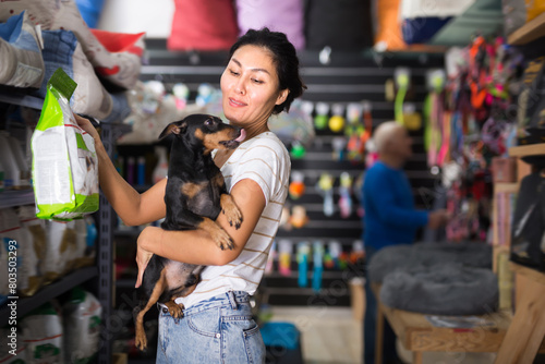 Asian woman with little dog in hands standing in salesroom of pet shop and choosing dog food.