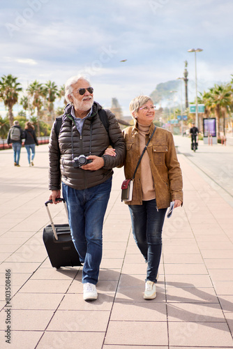 Wallpaper Mural Vertical. Couple of happy senior Caucasian tourist strolling hugging carefree with their luggage looking to the side. Pensioner people enjoying their journeys in a winter sunny day Torontodigital.ca