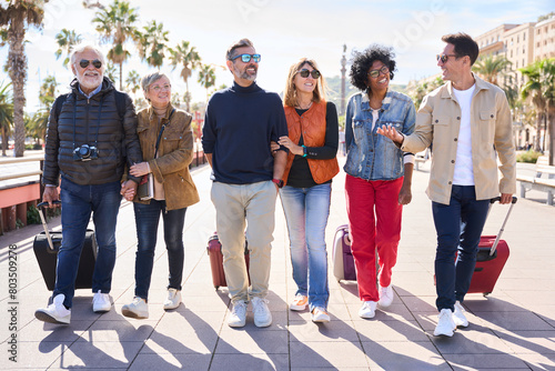 Group of smiling mature people walking carefree with their luggage enjoy vacations on street of Barcelona city. Diverse happy middle-aged friends celebrating leisure time on spring sunny day #803509278