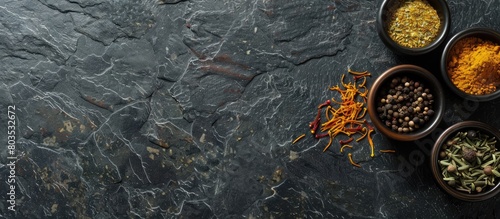 Assortment of spices including turmeric, saffron, dried thyme, cinnamon, nutmeg, and anise arranged in cups on a dark slate, stone, or concrete surface. Viewed from above with empty space for text.