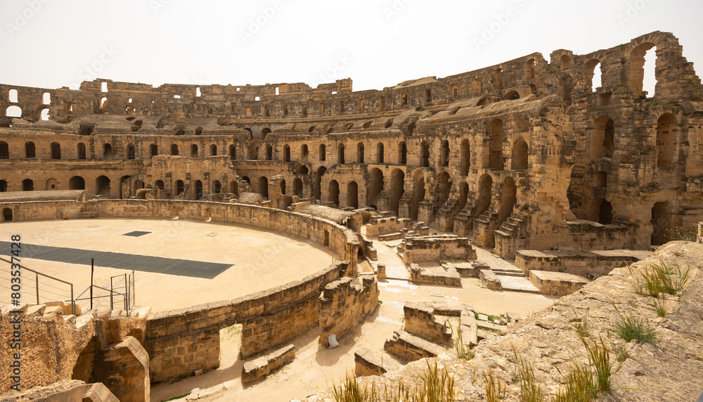 Scenic view of sun-drenched restored El Jem Amphitheatre arena encircled by ancient stone tiers and crumbling walls with archways under clear Tunisian sky