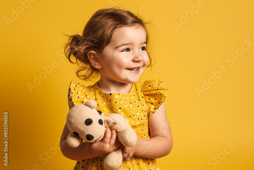 imagine A giggling toddler girl in a polka dot dress, holding a plush toy, standing against a sunny yellow backdrop.