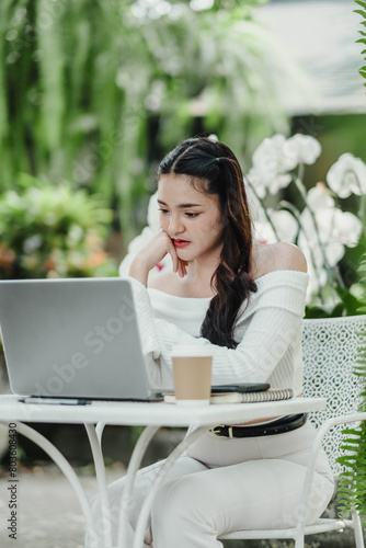 Young woman is deeply focused on her laptop screen while working in a lush garden setting with a coffee cup nearby.