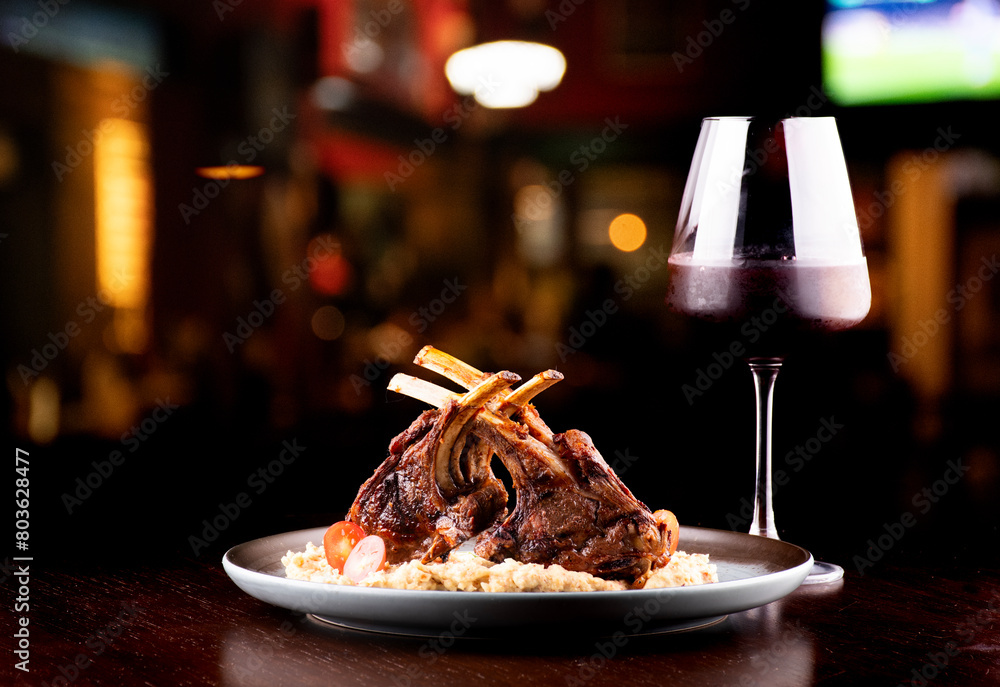 grilled rack of lamb with potatoes paired with glass of red wine on blurred pub background