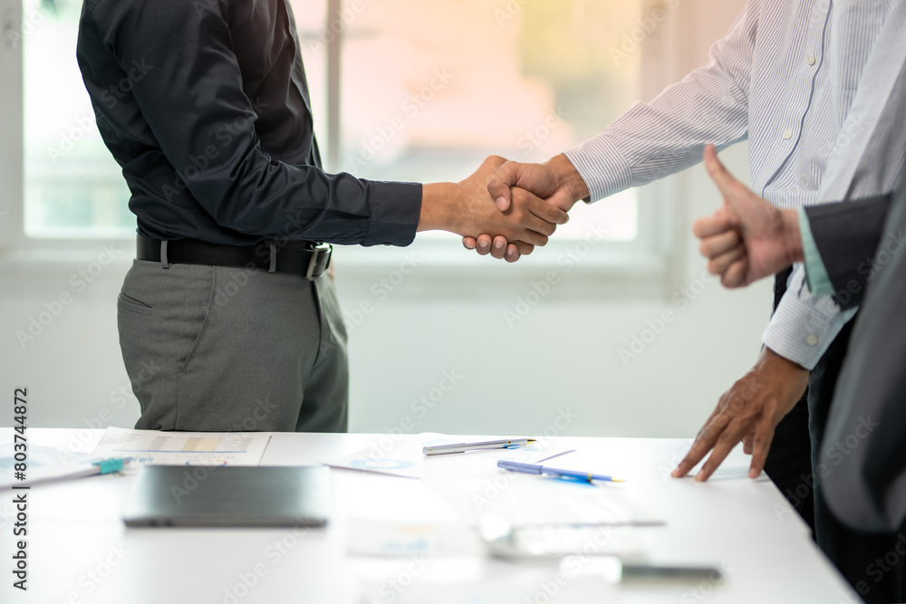 Three men shake hands in a business meeting. One of them is giving a thumbs up. The meeting is taking place in a conference room with a table and chairs Three men shake hands in a business meeting. One of them is giving a thumbs up. The meeting is taking place in a conference room with a table and chairs
