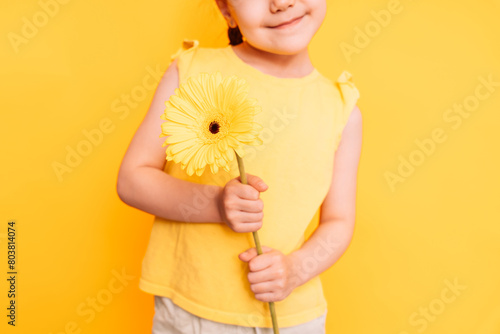 Little girl holding a yellow flower in front of a yellow background.