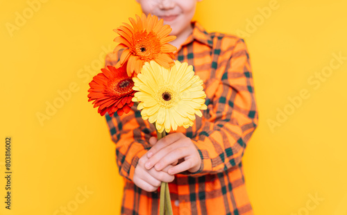 Boy in shirt holding flowers in front of him against yellow background.