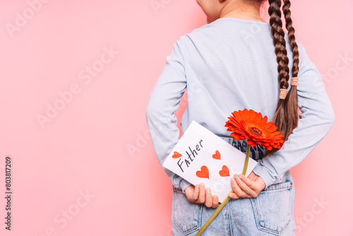 Little girl holding a drawn card and a flower behind her back for Father's Day holiday in front of a pink background