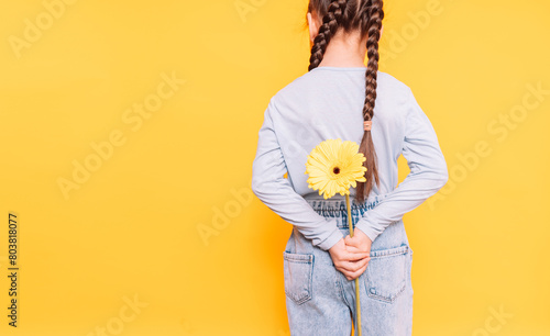 Child holding a yellow flower behind her back against a yellow background.
