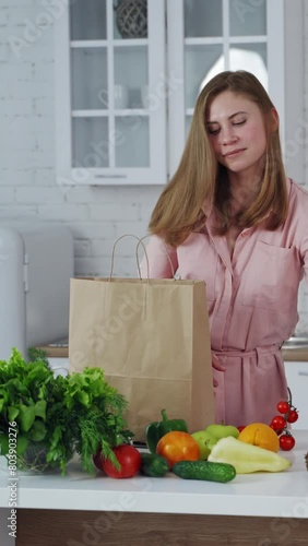 Happy housewife with fresh fruit and vegetables in the kitchen. Young woman unpack organic food from paper bag at home. Vertical video