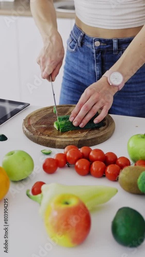 Delicious fruit and vegetables on a table and woman cooking. Housewife is cutting green cucumbers on a wooden board for making fresh salad in the kitchen. Vertical video