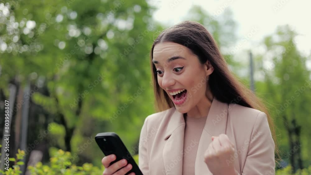Young attractive woman cheering emotionally while holding mobile telephone and going by green pedestrian area. Charming woman bending hand with clenched fist while winning prize in online lottery. Young attractive woman cheering emotionally while holding mobile telephone and going by green pedestrian area. Charming woman bending hand with clenched fist while winning prize in online lottery.