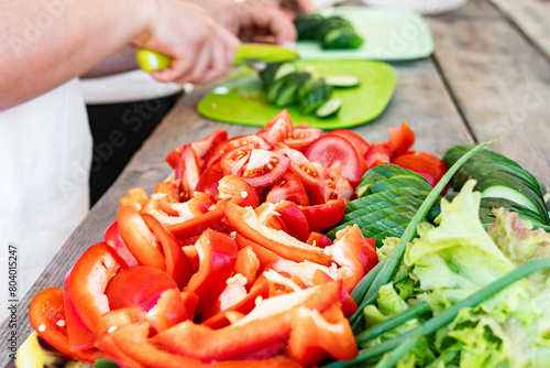 Sliced red bell peppers, tomatoes, and a variety of leafy greens and hands slicing cucumbers on green cutting boards on background