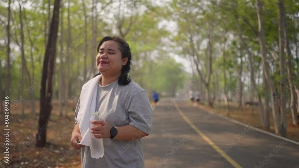 Tired overweight young Asian woman taking a water break from her morning walk at a public park Tired overweight young Asian woman taking a water break from her morning walk at a public park