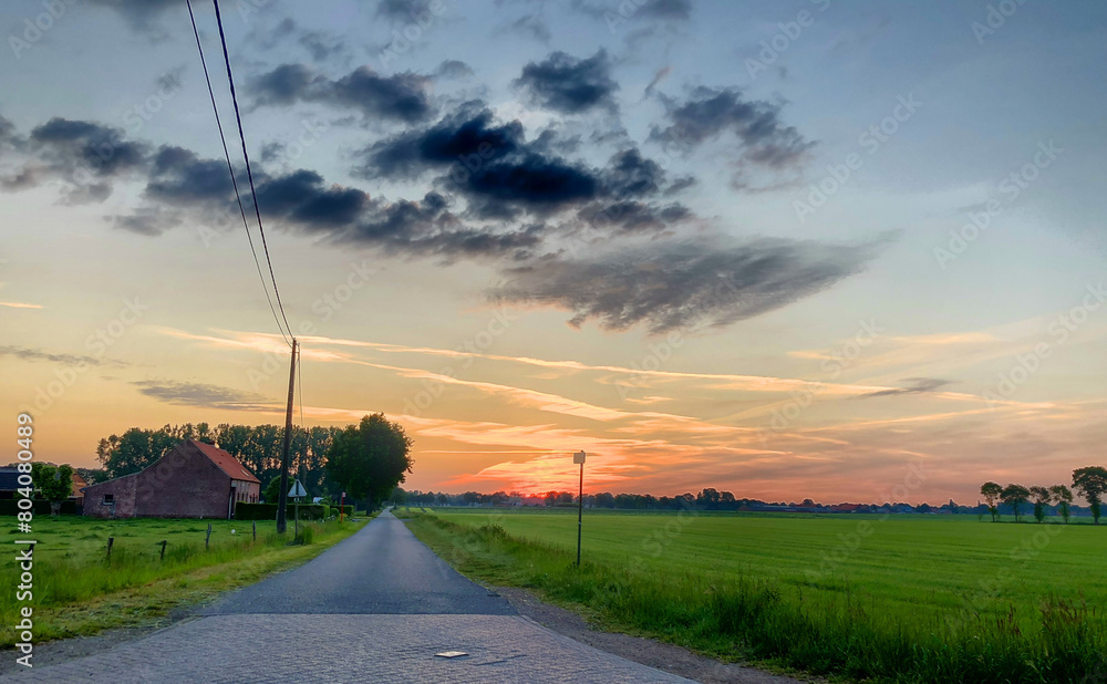 Fototapeta premium This image captures the breathtaking beauty of a sunrise over a rural landscape, featuring a long road leading past a traditional farmhouse surrounded by lush green fields. The sky, painted with