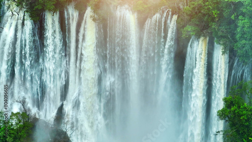 A hidden gem in the heart of a lush tropical rainforest, a colossal waterfall cascades through dense emerald foliage, unveiled by a soaring drone. Lush Woodlands: Primeval Forest. Thailand.
