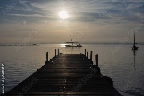 In the foreground  the wooden pier of the Albufera lake port is visible  with small boats sailing in the background