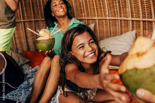 Happy girl taking coconut while sitting with family in wicker hut photo