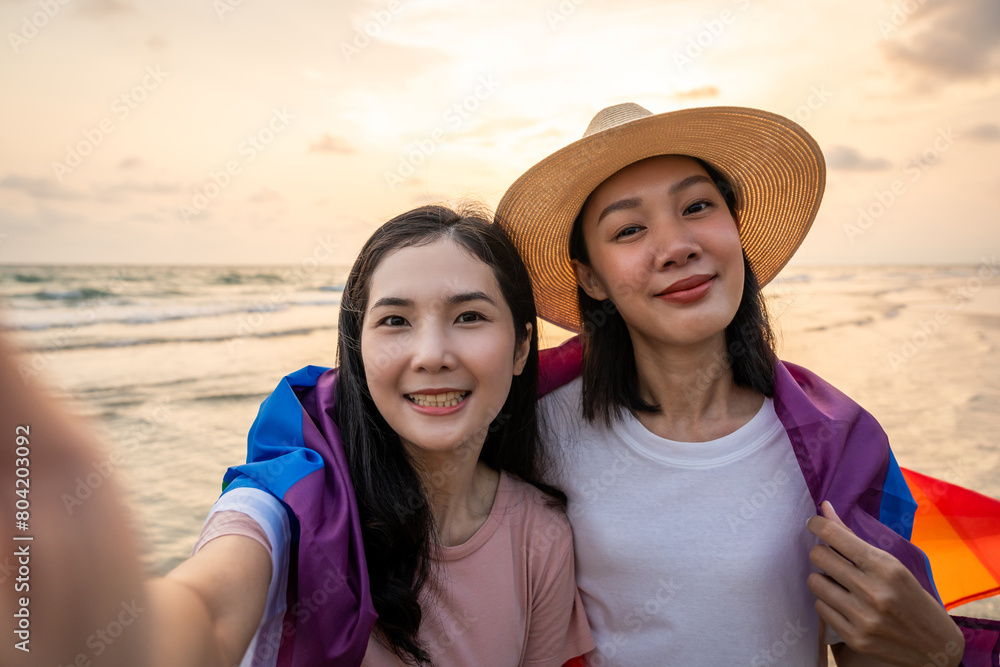 Young couple asian lesbian with pride movement LGBT holding rainbow flag for freedom. Demonstrate rights LGBTQ celebration pride Month lesbian Pride Symbol. Walking on the sand sea beach with sunset
