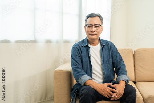 Handsome asian mature old man sitting on sofa couch in living room at home. Happy Portrait of cheerful smiling senior asian man relax in the house. Mature People and lifestyle