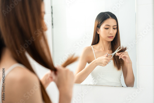 Young asian woman having problems with her hair Thin damaged dry hair falls out easily and is not healthy have to cut off. Standing looking mirror in restroom
